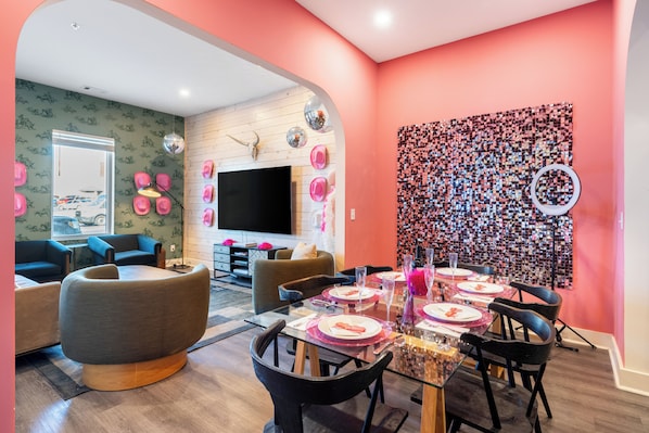A modern living room with a dining area, featuring a large flat-screen TV, comfortable seating, and a vibrant wall art piece.