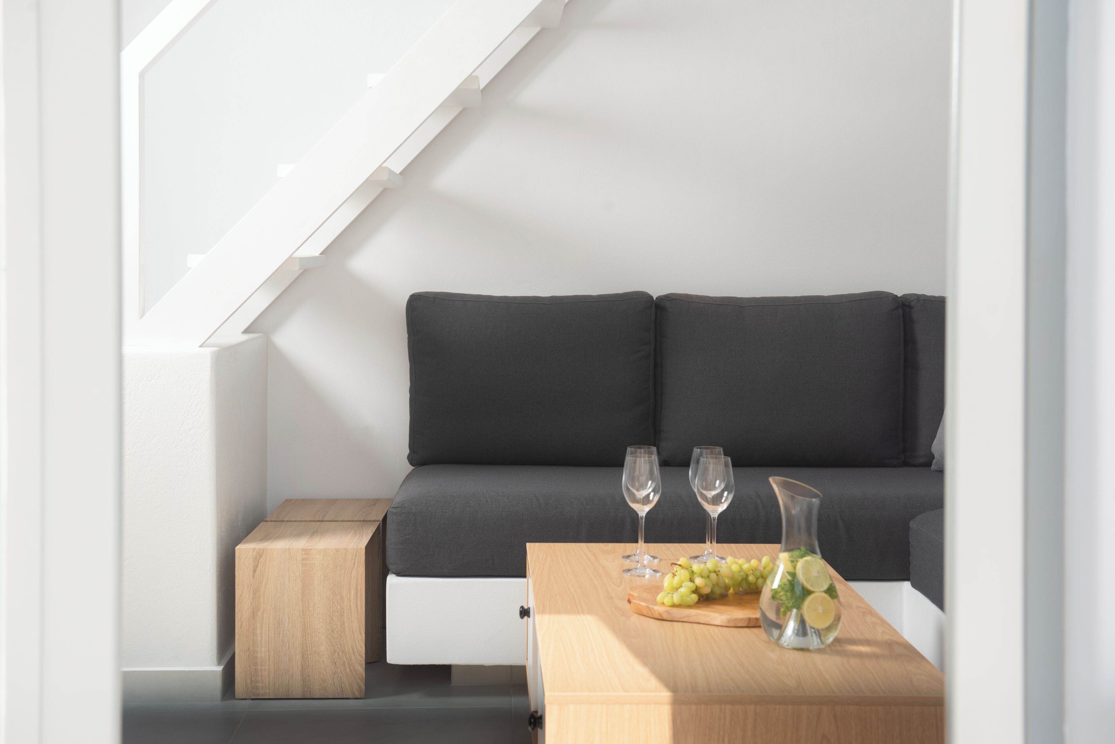A modern interior with a dark grey sofa, a wooden side table with a glass vase and grapes, and two wine glasses.