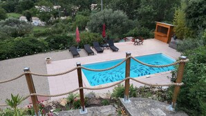 A swimming pool with a rope barrier, surrounded by lounge chairs and a small wooden structure, set in a landscaped garden with various plants and a view of a residential area.
