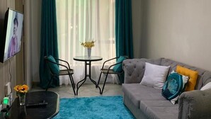 Un espace de vie compact avec un canapé gris, une petite table ronde et deux chaises. Un téléviseur est fixé au mur, et un luminaire au plafond est visible.