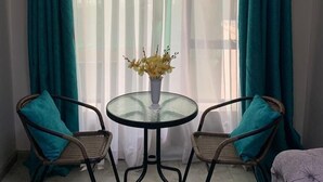 Une pièce avec une table en verre, deux chaises en osier, des rideaux turquoise et un vase rempli de fleurs jaunes.