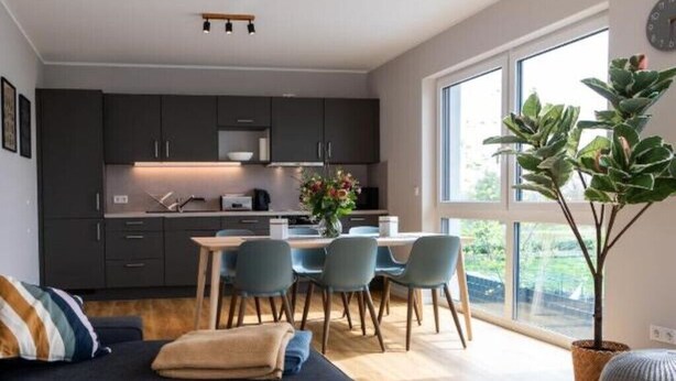 A modern kitchen with dark cabinets, a dining area with a table and chairs, and a large window offering a view of greenery. - Moderne & Familienfreundliche Ferienwohnung am Goitzschesee (Muldestausee)