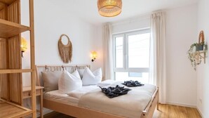 A bedroom with a wooden bed, white bedding, a woven headboard, a large window with curtains, and a wall-mounted plant. - Moderne & Familienfreundliche Ferienwohnung am Goitzschesee (Muldestausee)
