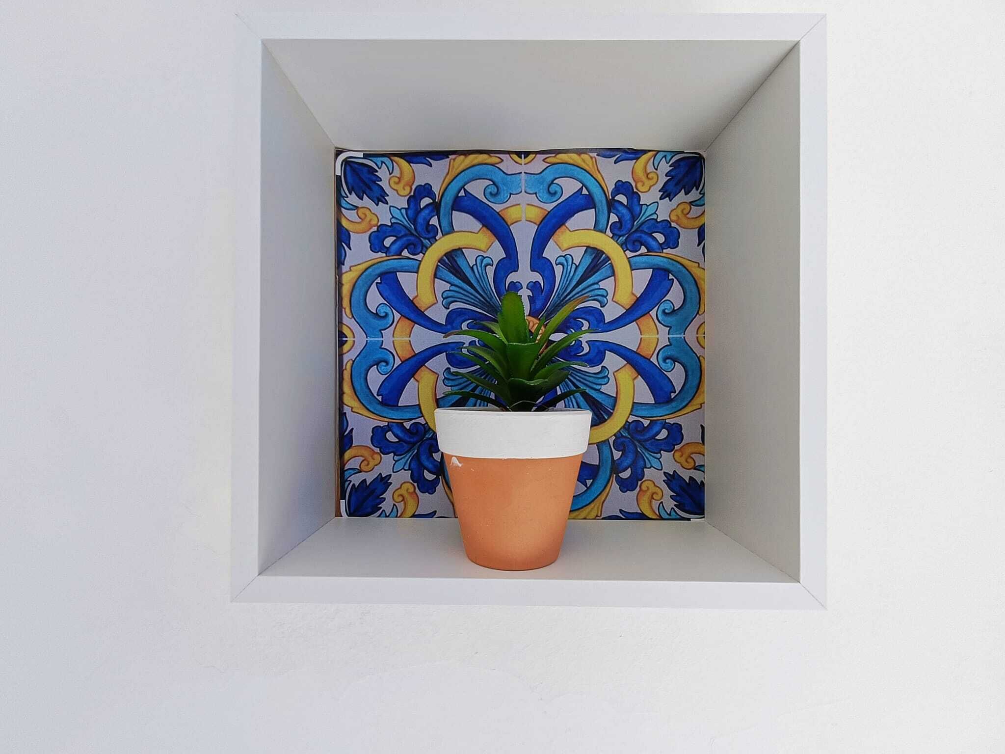 Ein Topfpflanze in einer weißen Pflanzkasten vor einer gefliesten Wand mit blau-gelbem Muster.