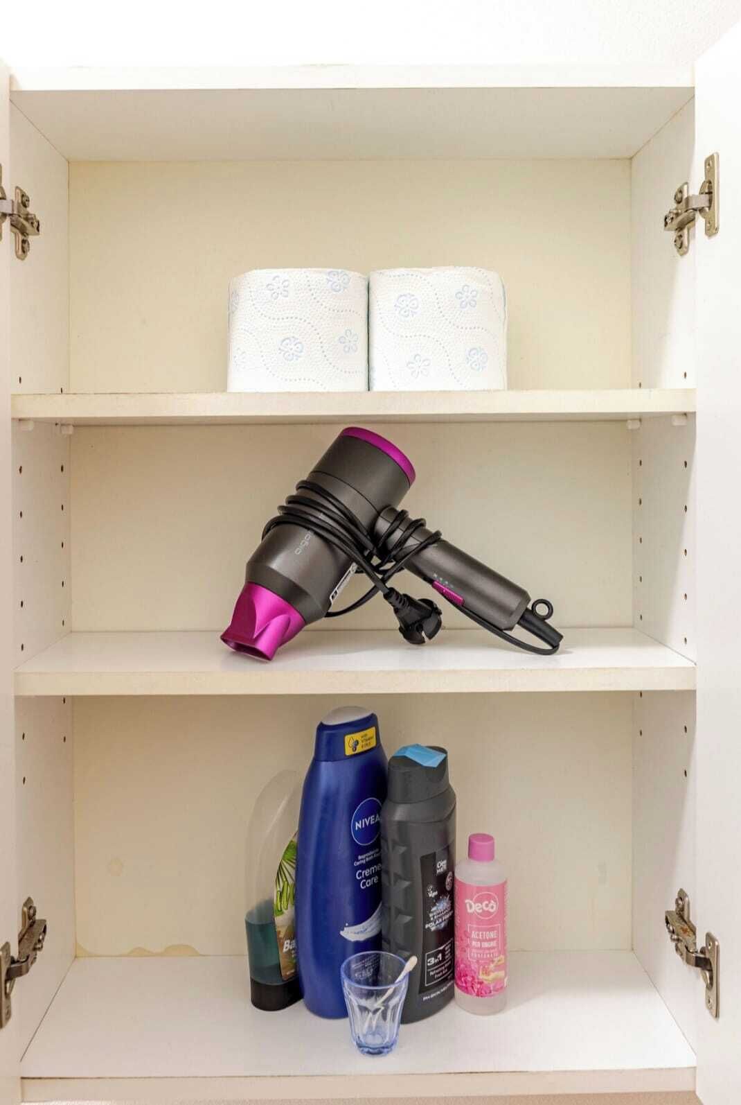 Een plank met een haardroger, twee rollen toiletpapier, een blauwe fles Nivea crème, een zwarte fles aceton, een klein glas en een roze fles Deco aceton.