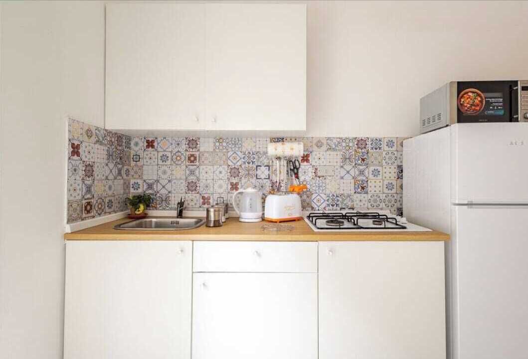 Una cocina con armarios blancos, salpicadero de azulejos, estufa a gas, microondas y un lavamanos.