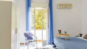 A bedroom with a bed, a chair, a small table with a cup and flowers, and a window with curtains. - Appartamenti Abate Riva del Golfo Home (Alcamo)