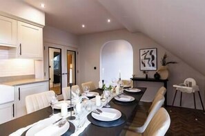 Un espace repas avec une longue table dressée pour le repas, un coin cuisine avec des meubles blancs et une vue sur l’extérieur par une porte en arc de cercle.