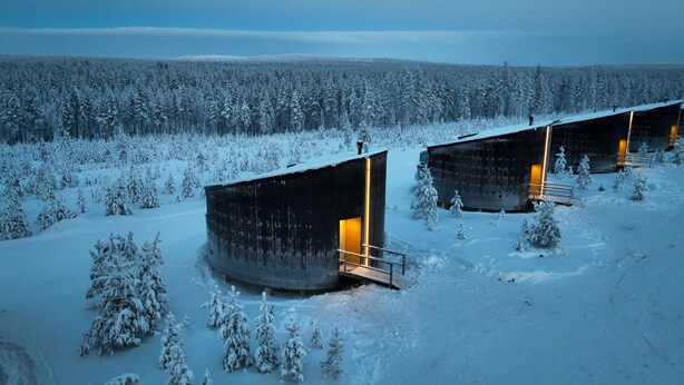 A snow-covered landscape with two rustic cabins, each featuring a small porch and a single illuminated window. - Livo Arctic (Posio)
