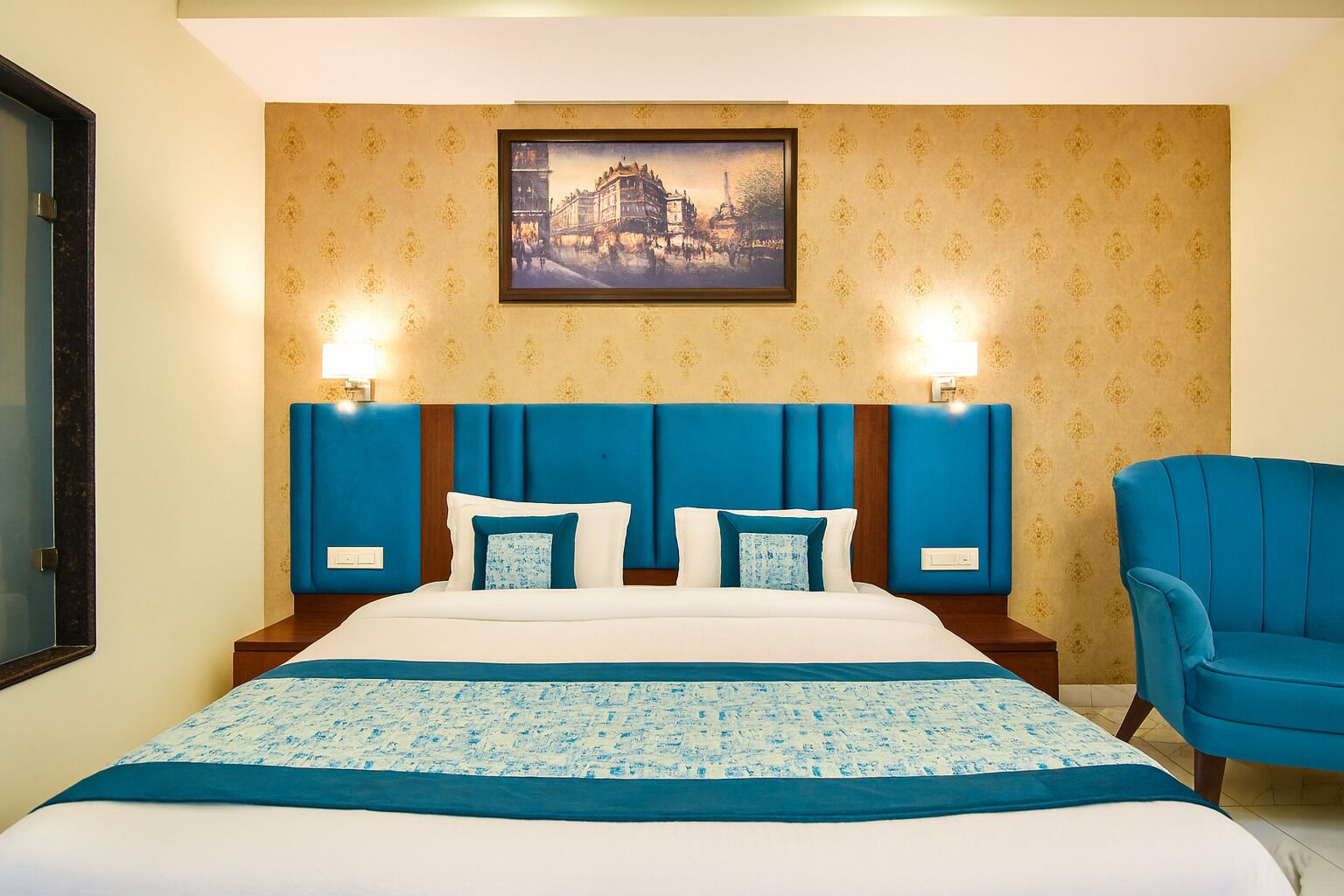 A hotel room with a large bed, two wall-mounted lamps, a blue armchair, and a framed picture on the wall.
