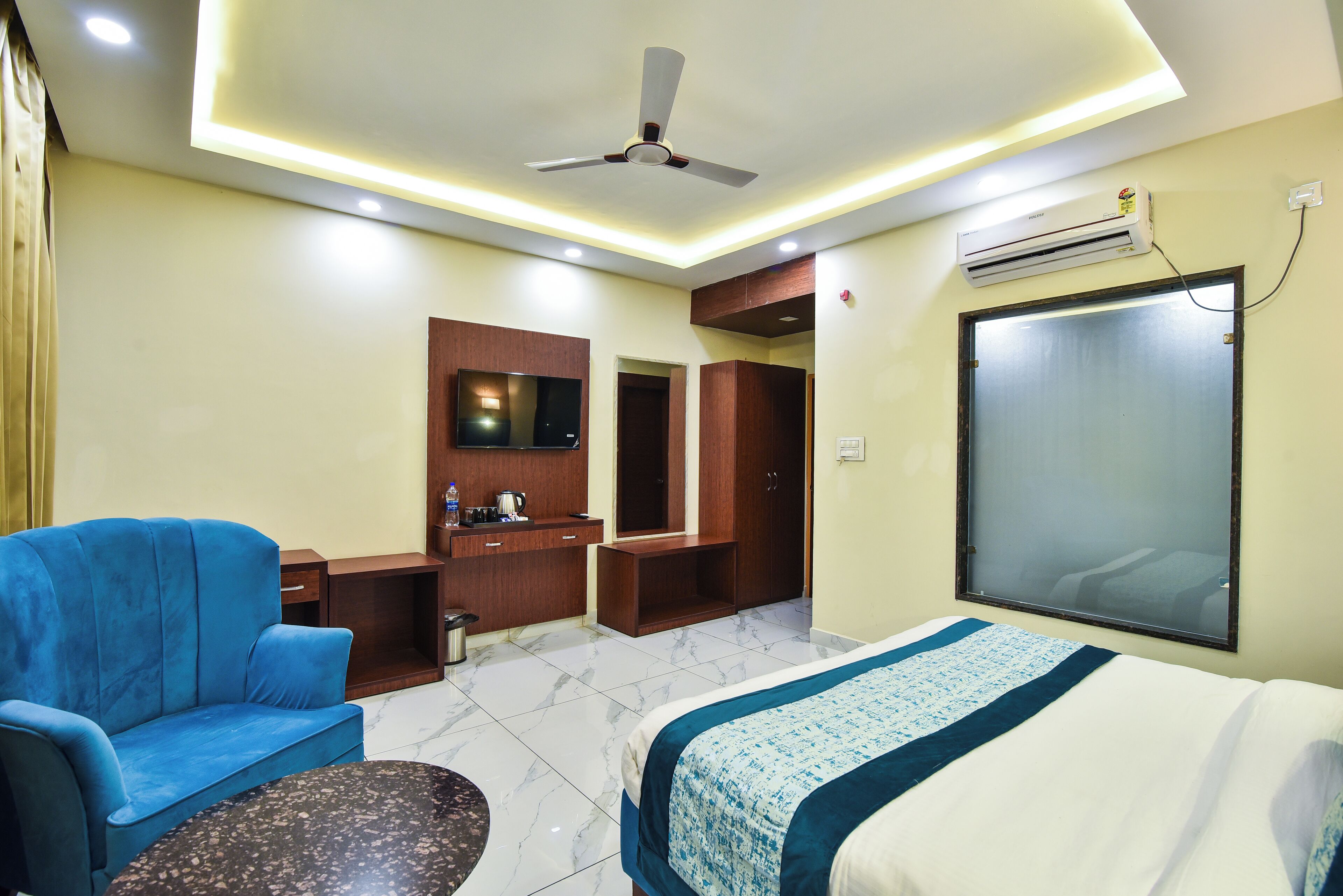 A hotel room with a bed, a blue sofa, a small table, a desk, a mirror, and an air conditioning unit.