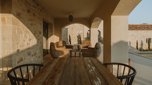 Een overdekt terras met stenen muren, rieten meubels en een houten tafel.