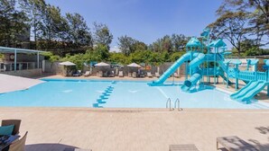 Una piscina con un tobogán azul y zona de juegos, rodeada de sillas de descanso y sombrillas.