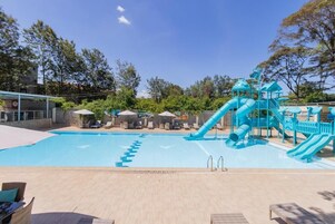 Una piscina con un tobogán azul y zona de juegos, rodeada de sillas de descanso y sombrillas.