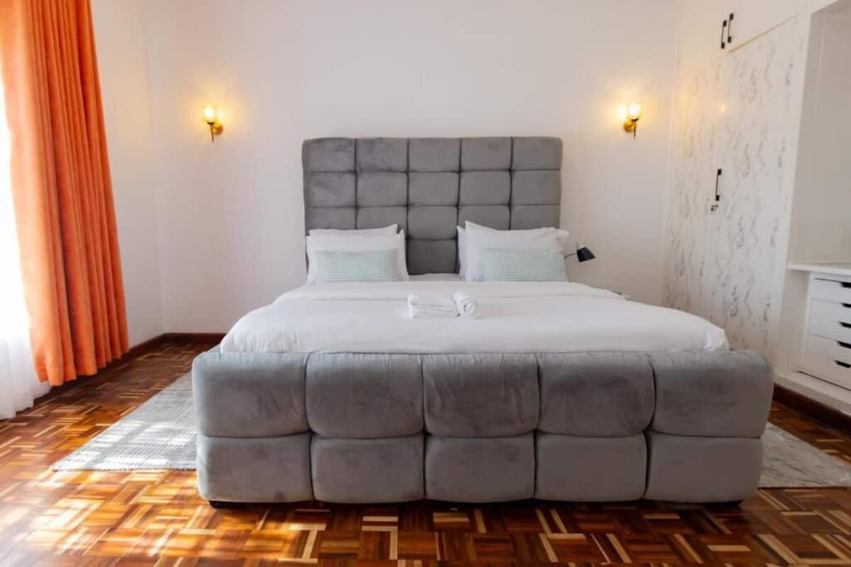 Une chambre à coucher avec une tête de lit capitonnée, un grand lit recouvert de draps blancs, deux lampes fixées au mur et un plancher en parquet.