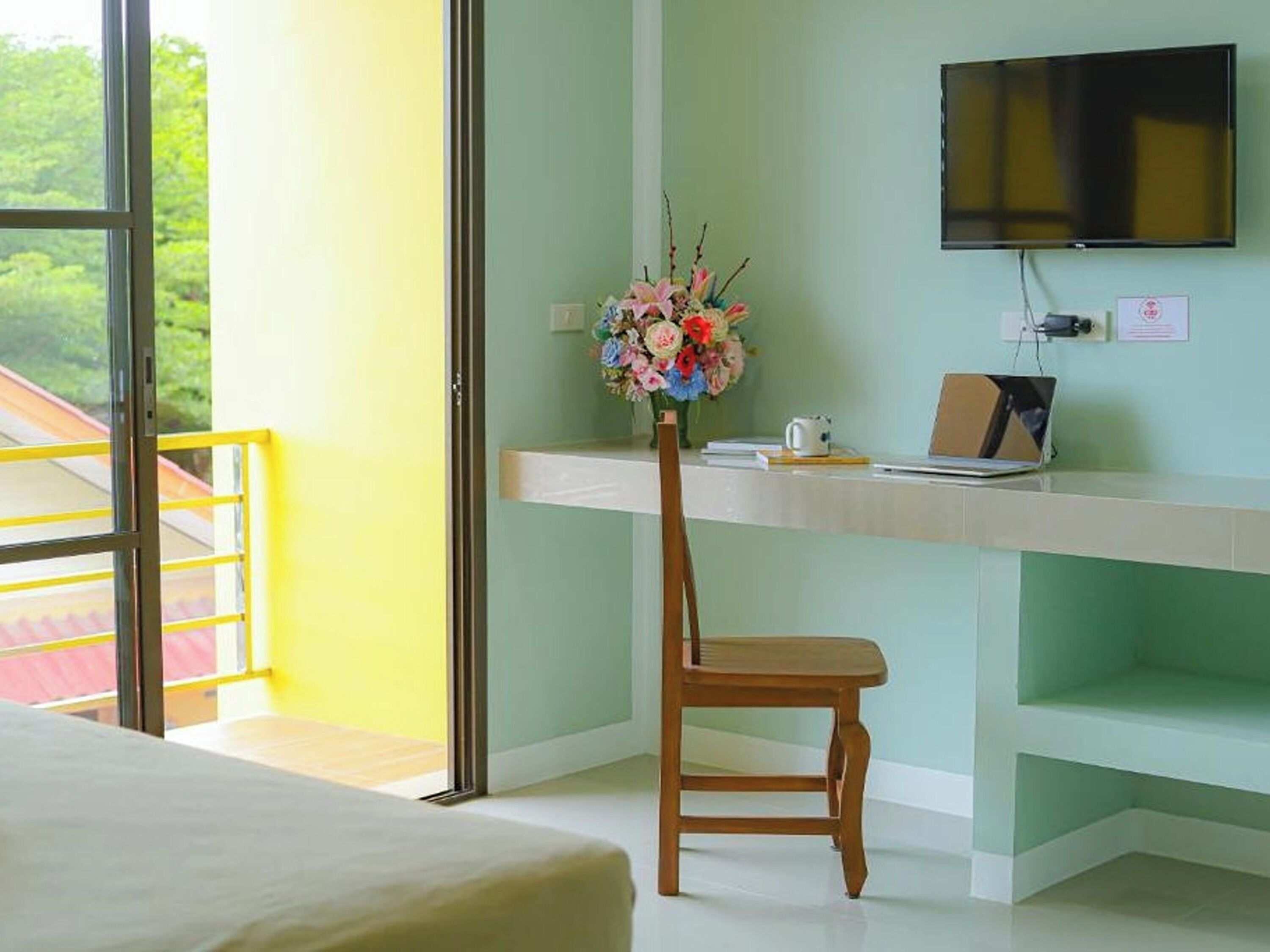 A room with a bed, a desk with a laptop, a TV, and a flower vase.
