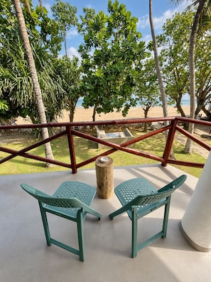 Un balcón con dos sillas de plástico verde, un taburete de madera y vistas a una playa con palmeras.