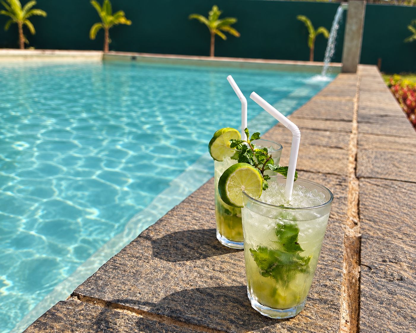 Two glasses of a beverage with lime and mint garnish on a stone ledge by a pool with clear water and palm trees in the background.