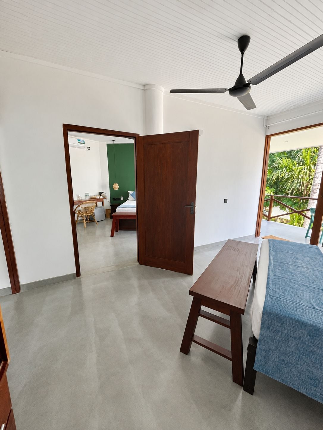 Una stanza con un letto, un tavolo di legno, un ventilatore a soffitto e vista su un balcone con piante.