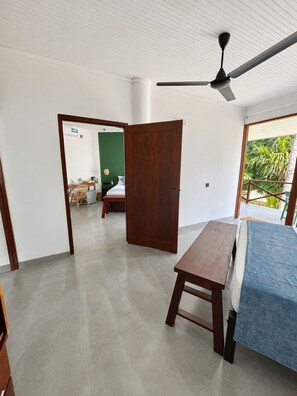 A room with a bed, a wooden table, a ceiling fan, and a view of a balcony with greenery. - Casa Smeralda (Batigama)