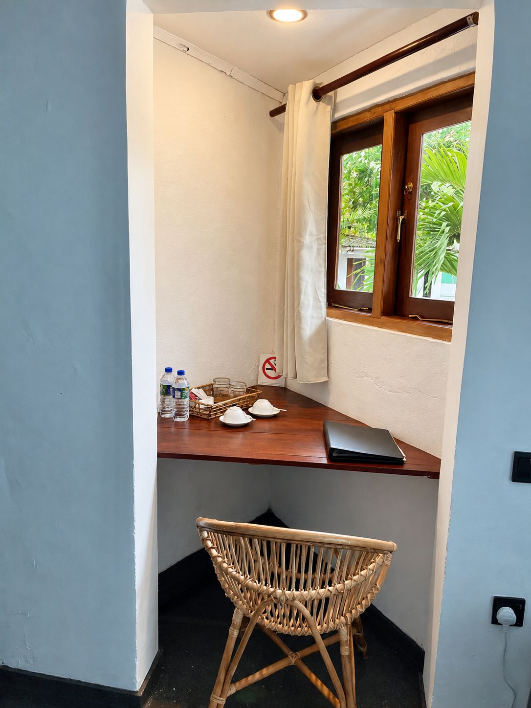 Een kleine kamer met een houten bureau, een rieten stoel, een raam met gordijn en een bordje ‘niet roken’.