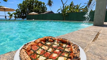 Pizza auf einem weißen Teller, neben einem Pool mit klarem, blauem Wasser und Palmen im Hintergrund.