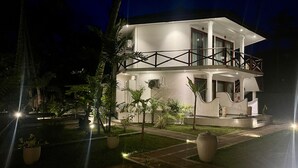 Un edificio de dos pisos con un balcón, rodeado de plantas en macetas y un jardín iluminado por la noche.