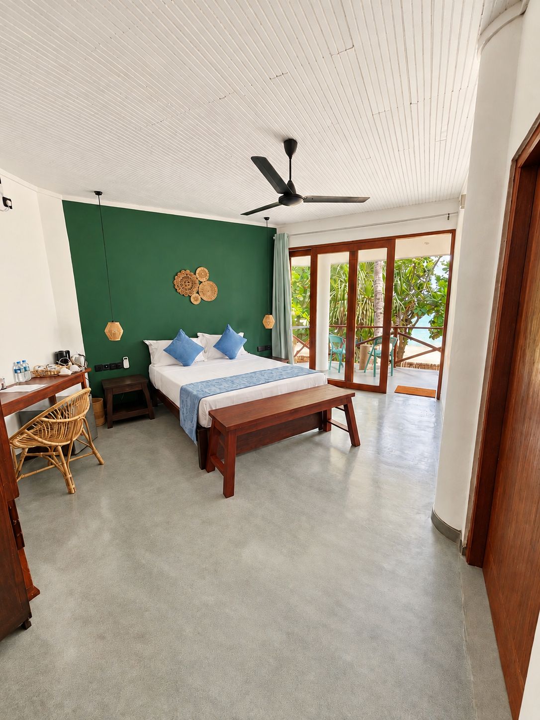 A bedroom with a bed, bedside tables, a desk, and a chair. There is a ceiling fan and a view of the outdoors through a sliding door.