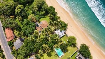 An aerial view of a coastal property with a swimming pool, beach access, and lush greenery.
