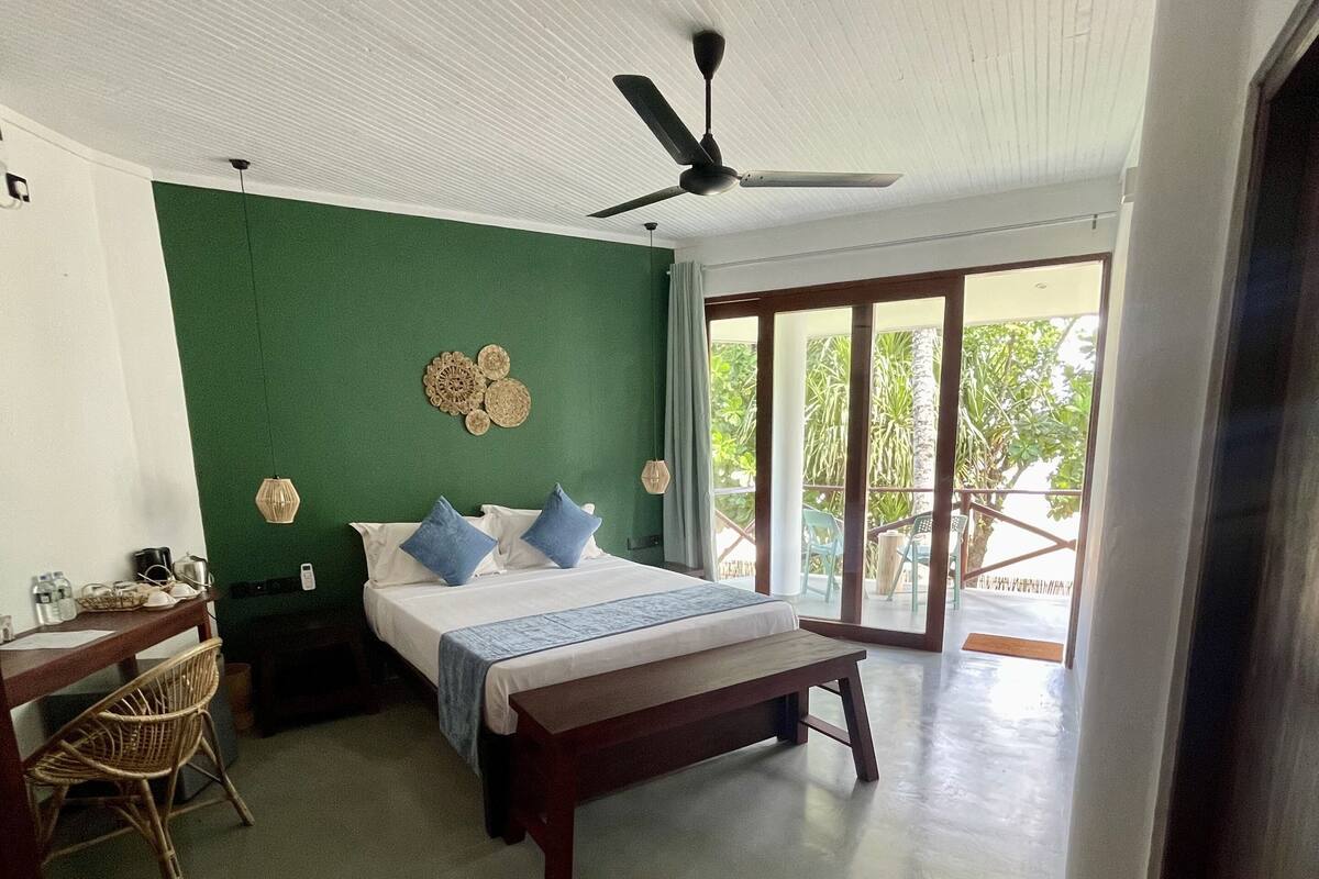 A bedroom with a bed, a desk, a chair, a ceiling fan, and a view of the outdoors.