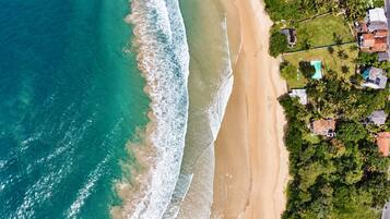 An aerial view of a beach with clear turquoise waters, a sandy shore, and a residential area with houses and greenery.
