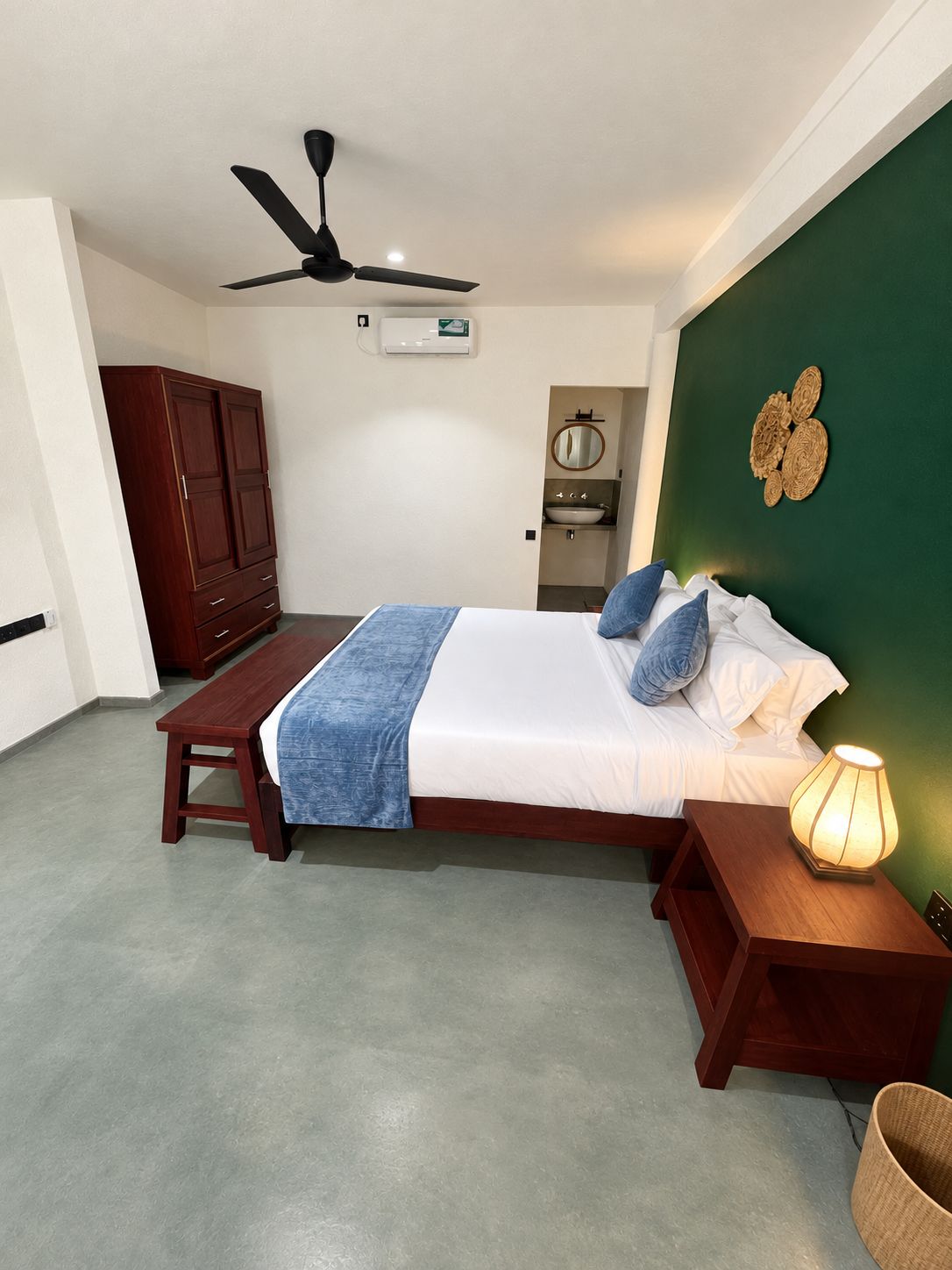 Une chambre à coucher comprenant un lit, une table de chevet avec une lampe, un ventilateur de plafond, une armoire et une petite salle de bain avec un lavabo et un miroir.