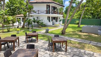 A two-story building with a balcony, surrounded by a garden with palm trees and a patio area with tables and chairs.