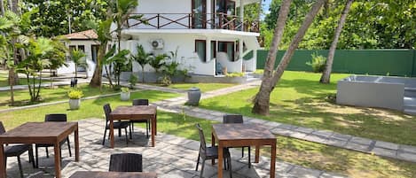 Un edificio de dos plantas con un balcón, rodeado por un jardín con palmeras y un patio con mesas y sillas.