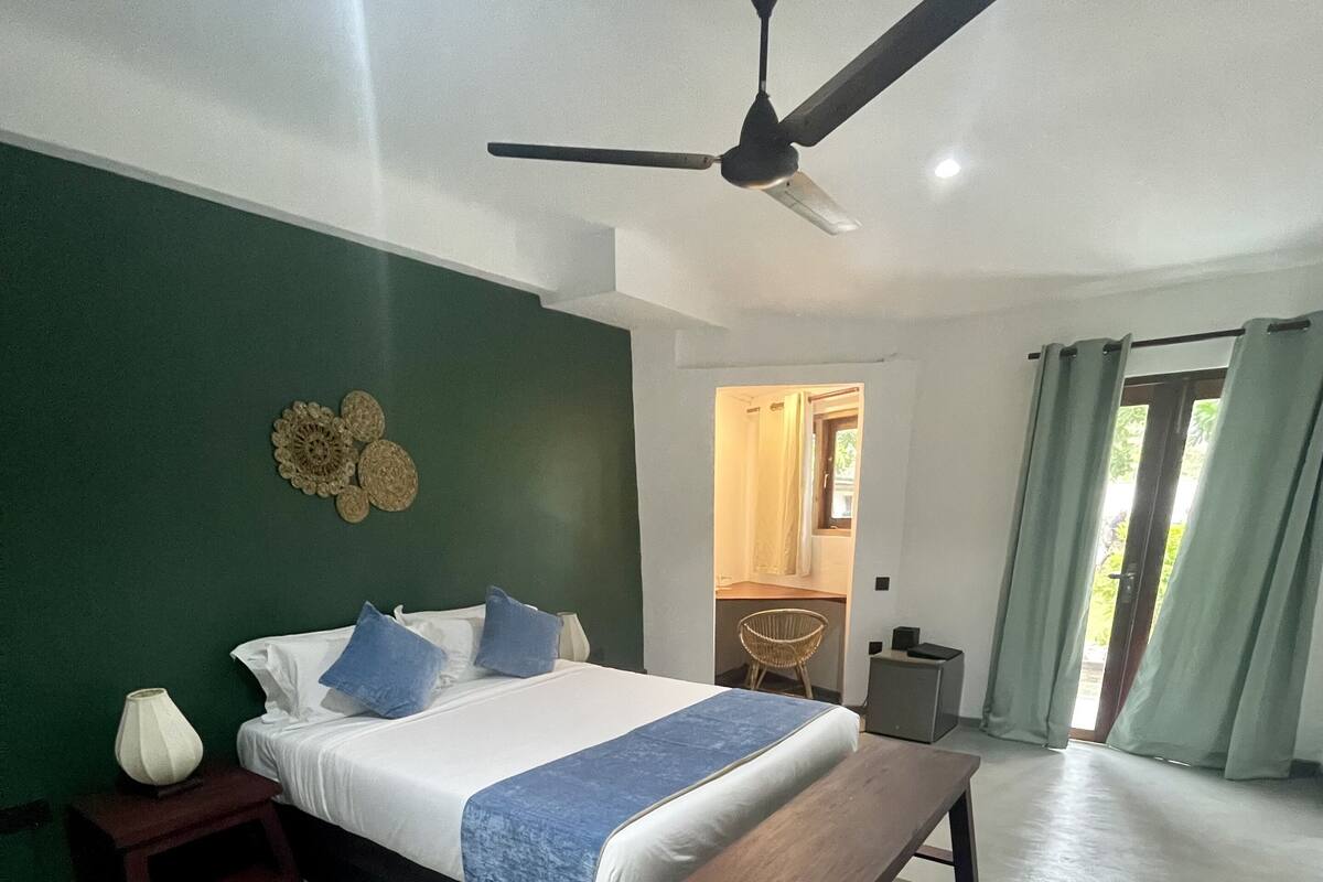 A bedroom with a bed, bedside tables, a bench, a ceiling fan, and a view of the outside.