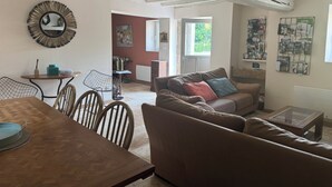 Uma sala de estar com uma mesa de jantar de madeira, um sofá com almofadas, uma cadeira de metal e um espelho fixado na parede.