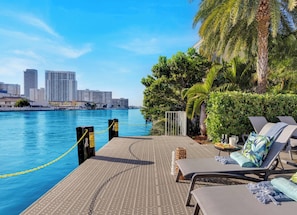 Un salón junto al agua con sillones de sol, una mesa junto a la piscina y un horizonte urbano al fondo.