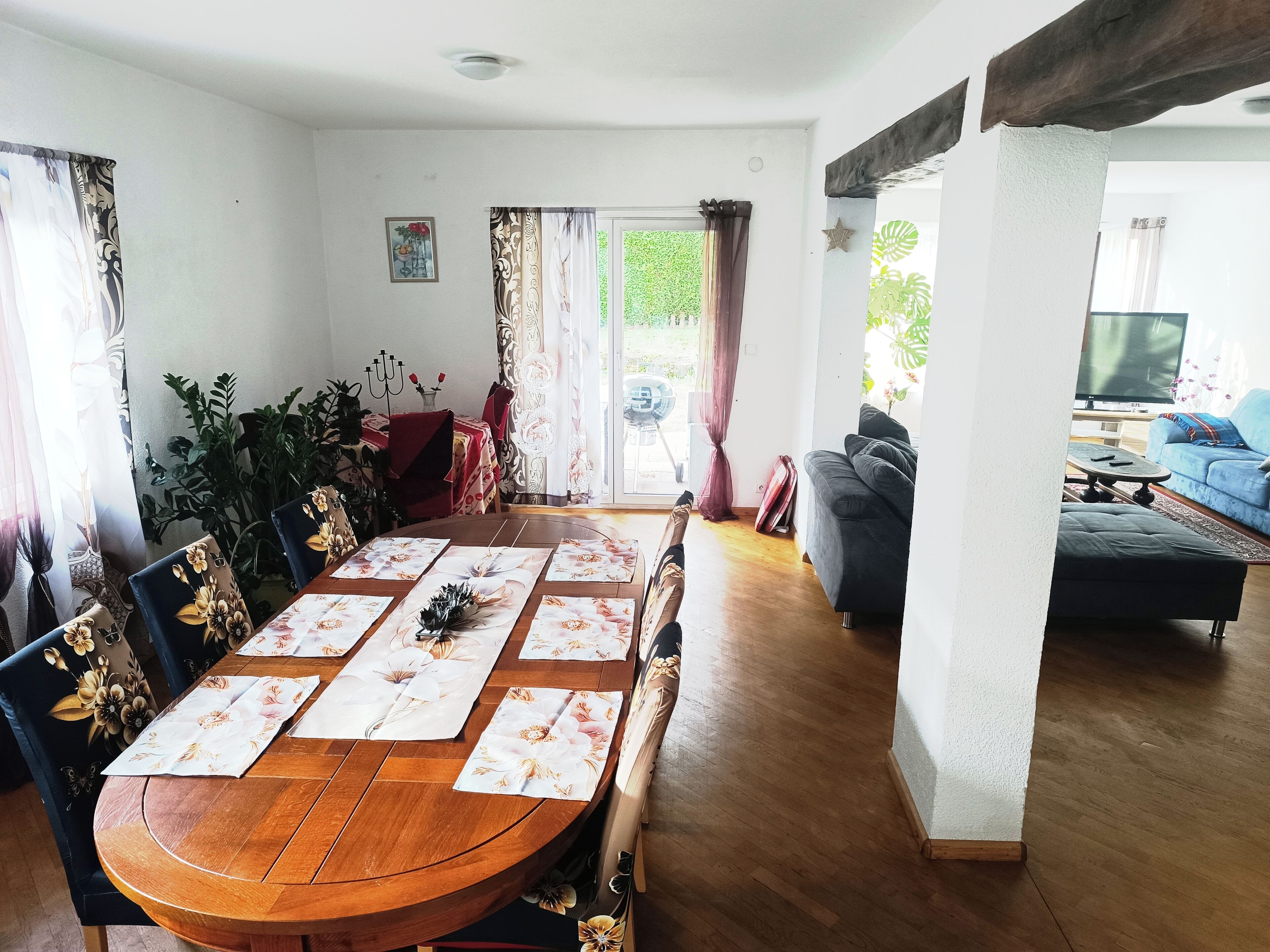 A dining area with a wooden table set for four, surrounded by chairs with floral upholstery. A dining table with a red tablecloth and a vase of flowers is visible. The room has a wooden floor and a sofa in the background.