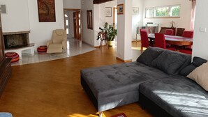 Un vaste salon comprenant un canapé d’angle gris, un fauteuil beige et une cheminée. On y trouve également un coin repas avec des chaises rouges et une table, ainsi qu’une cuisine à l’arrière-plan.