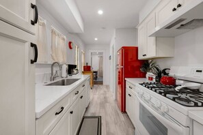 Une cuisine étroite avec des meubles blancs, un réfrigérateur rouge, une cuisinière à gaz et une machine à café.