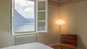 Una habitación de hotel con cama, sillón, ventana y vista a un lago y montañas, además de una lámpara.