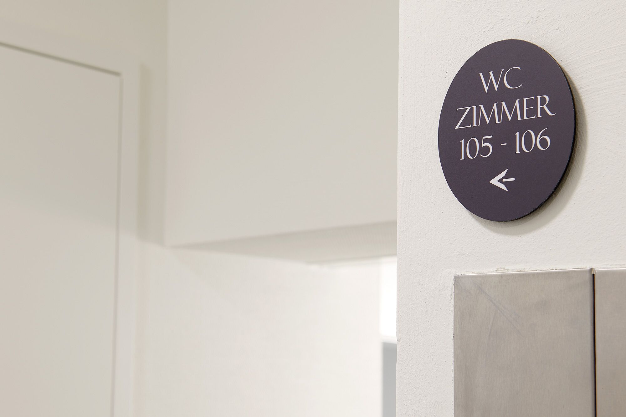 Un letrero en la pared indica los servicios sanitarios y las habitaciones Zimmer, números 105-106, con una flecha que apunta a la izquierda.