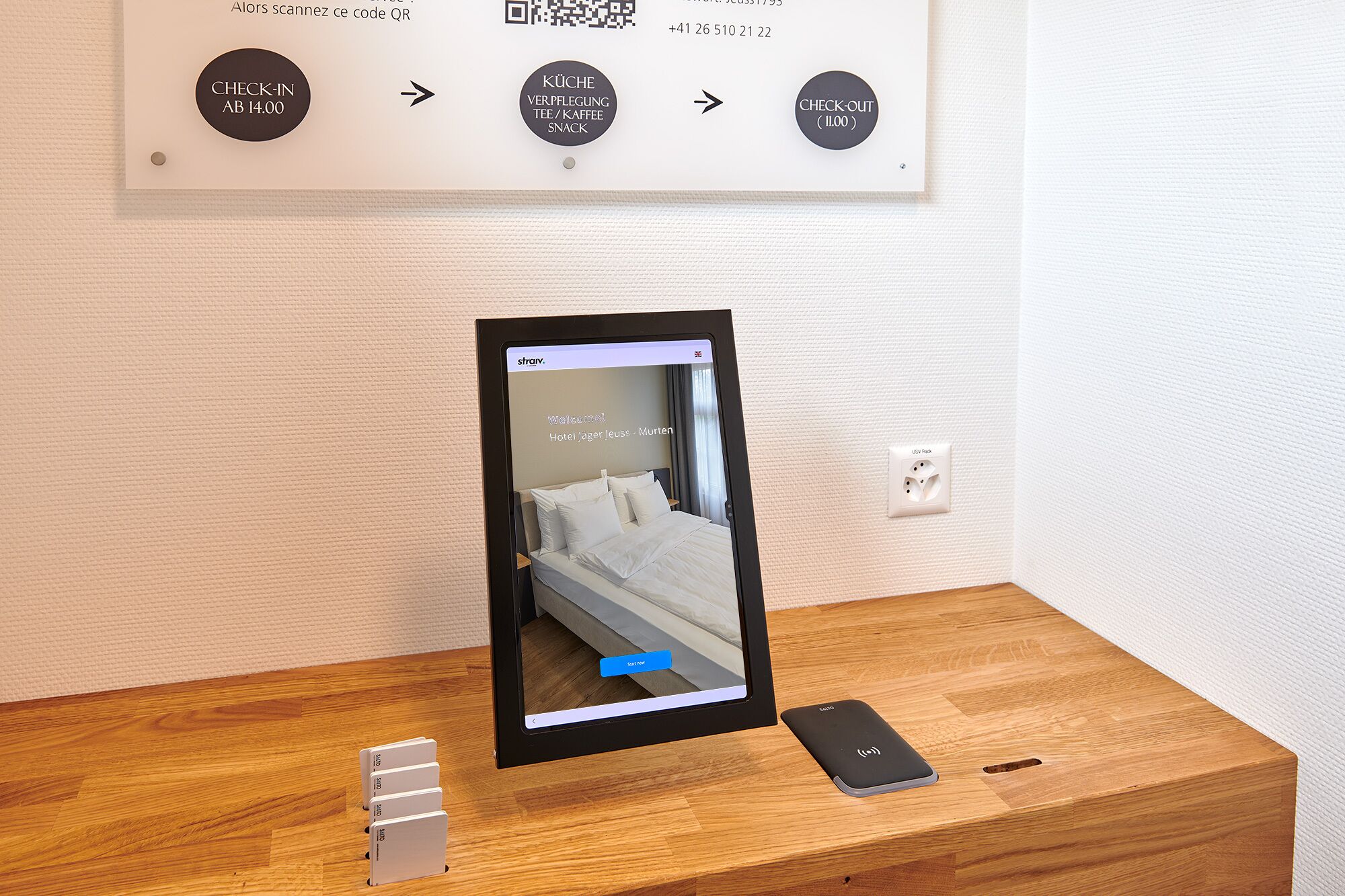 A hotel room with a bed, a bedside table, a charging station, and a QR code for check-in and check-out.