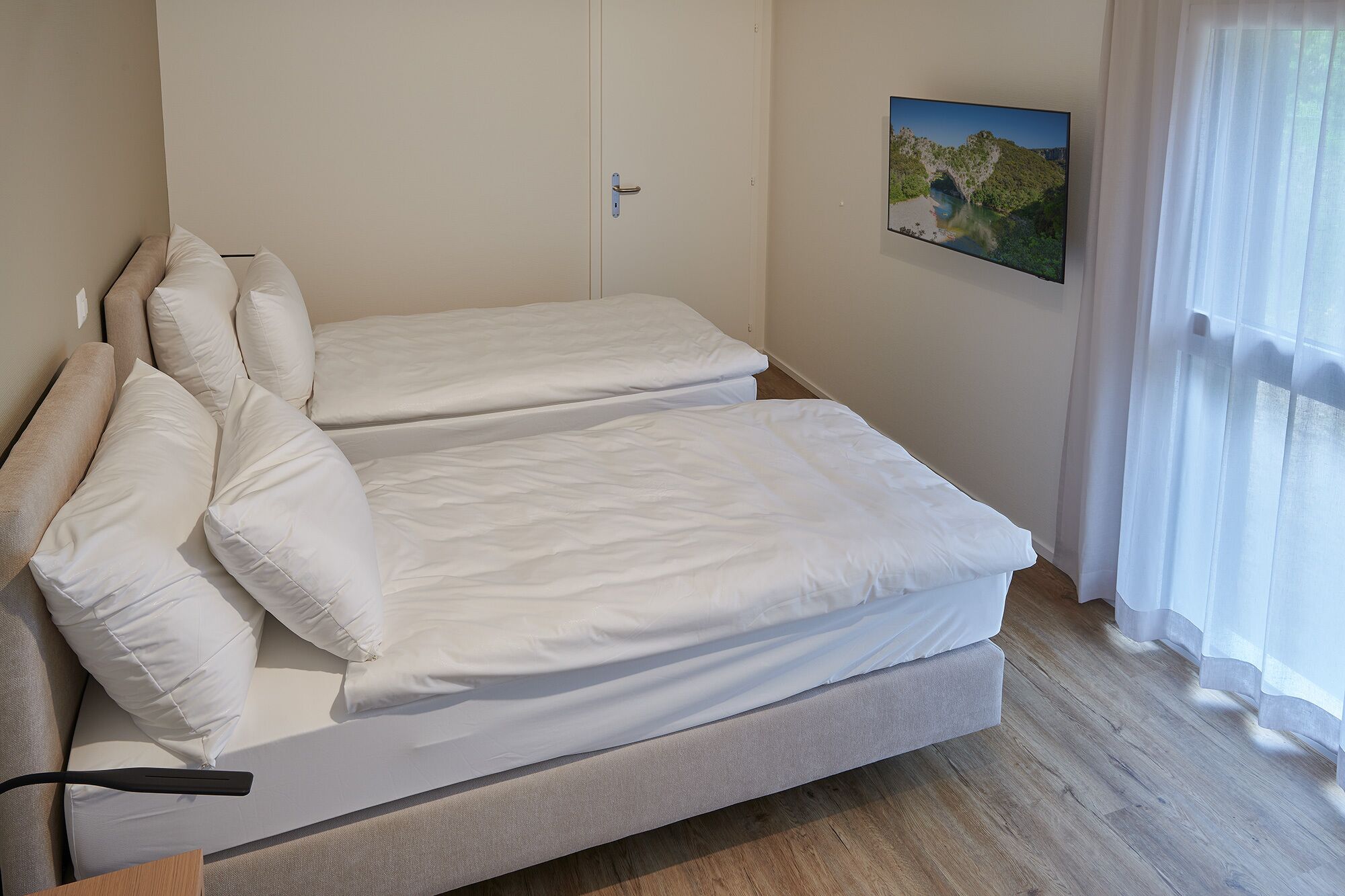 A bedroom with two single beds, a wooden floor, a door, and a window with curtains.