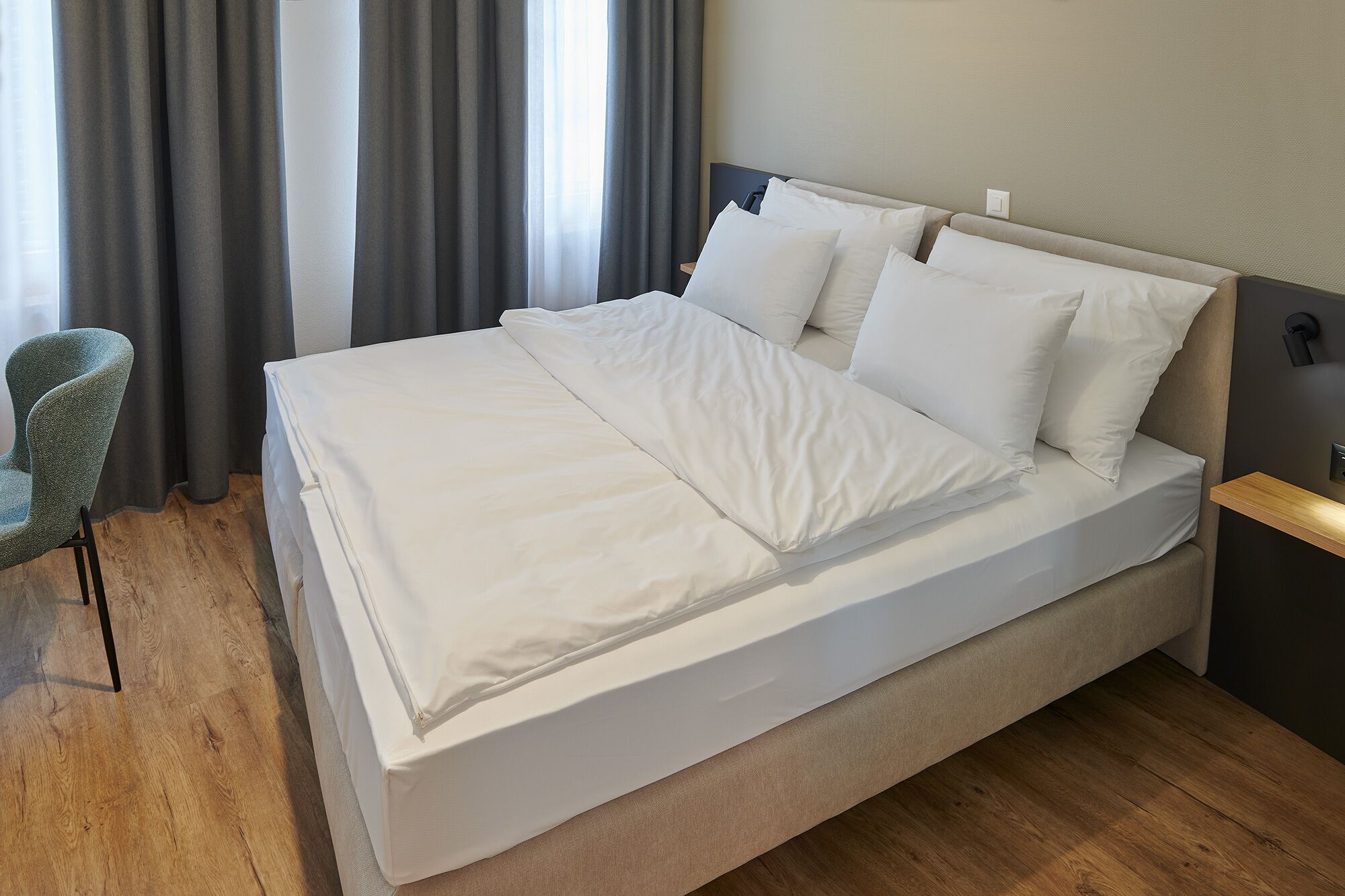 A neatly made bed with white linens in a room with wooden flooring and a grey chair.