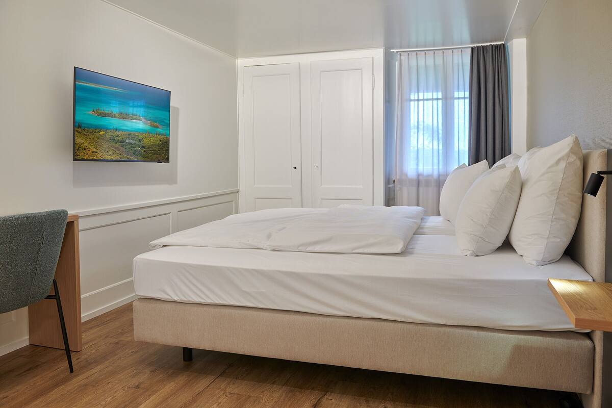 A bedroom with a bed, a nightstand, a chair, a painting, and a window with curtains.