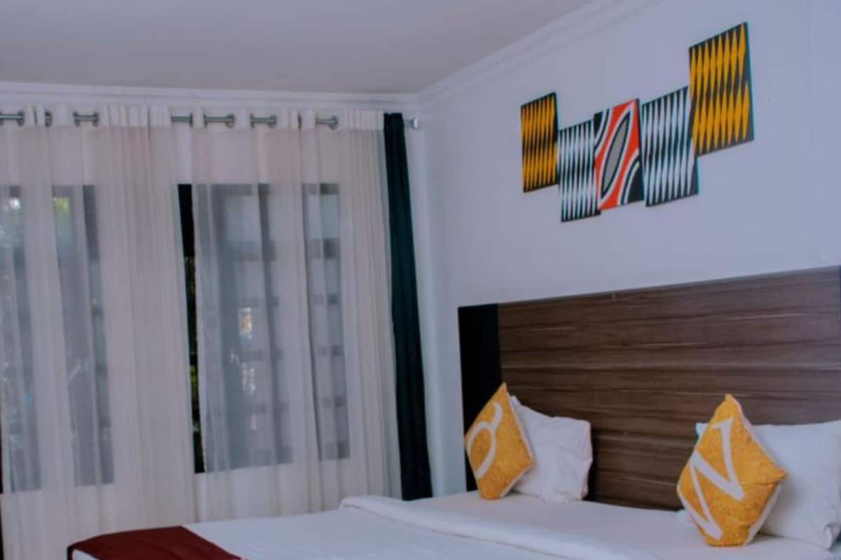 A hotel room with a large bed, white bedding, and a red throw blanket. There is a wooden headboard and two decorative pillows.