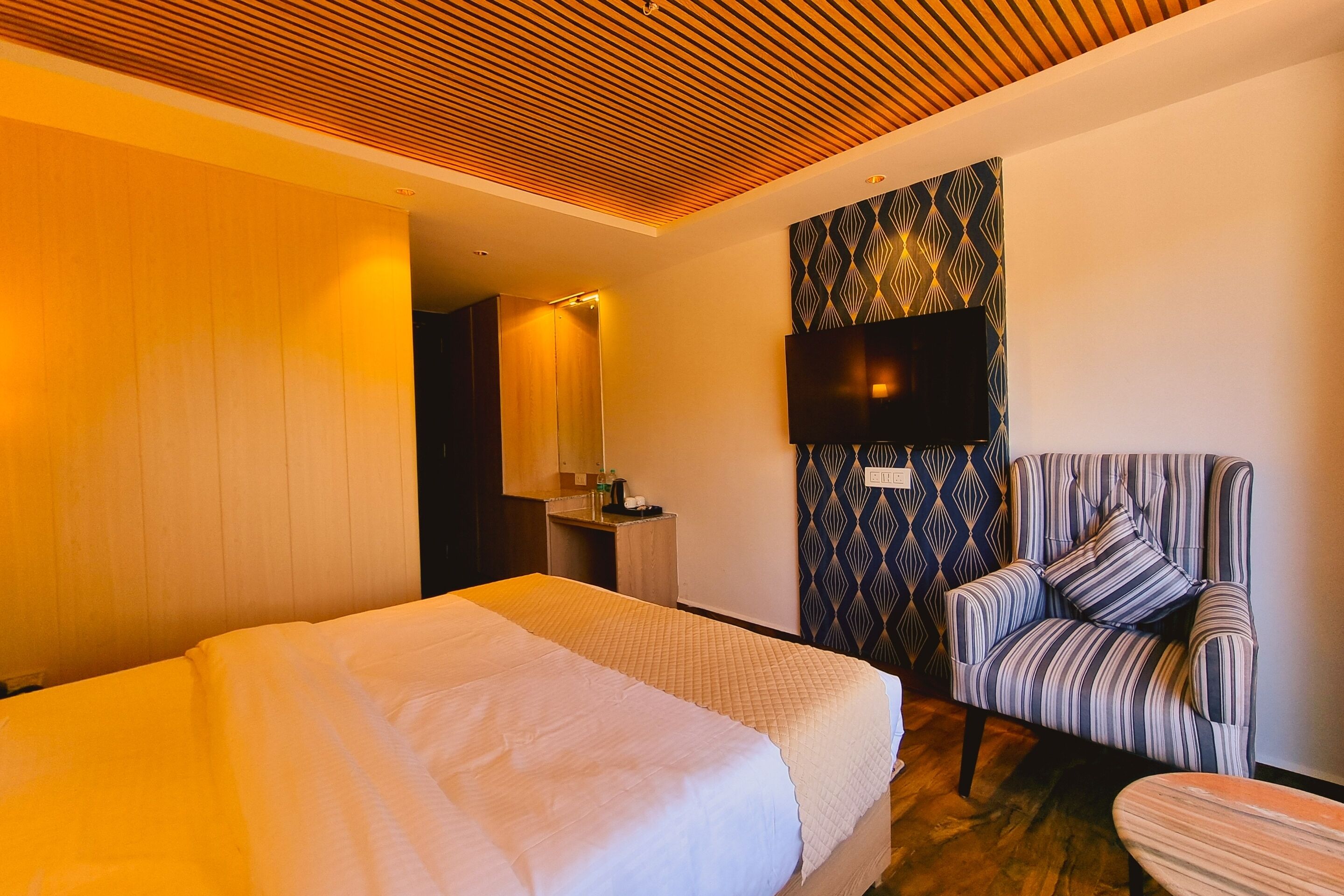 A hotel room with a bed, a striped armchair, a TV mounted on the wall, and a patterned wallpaper.