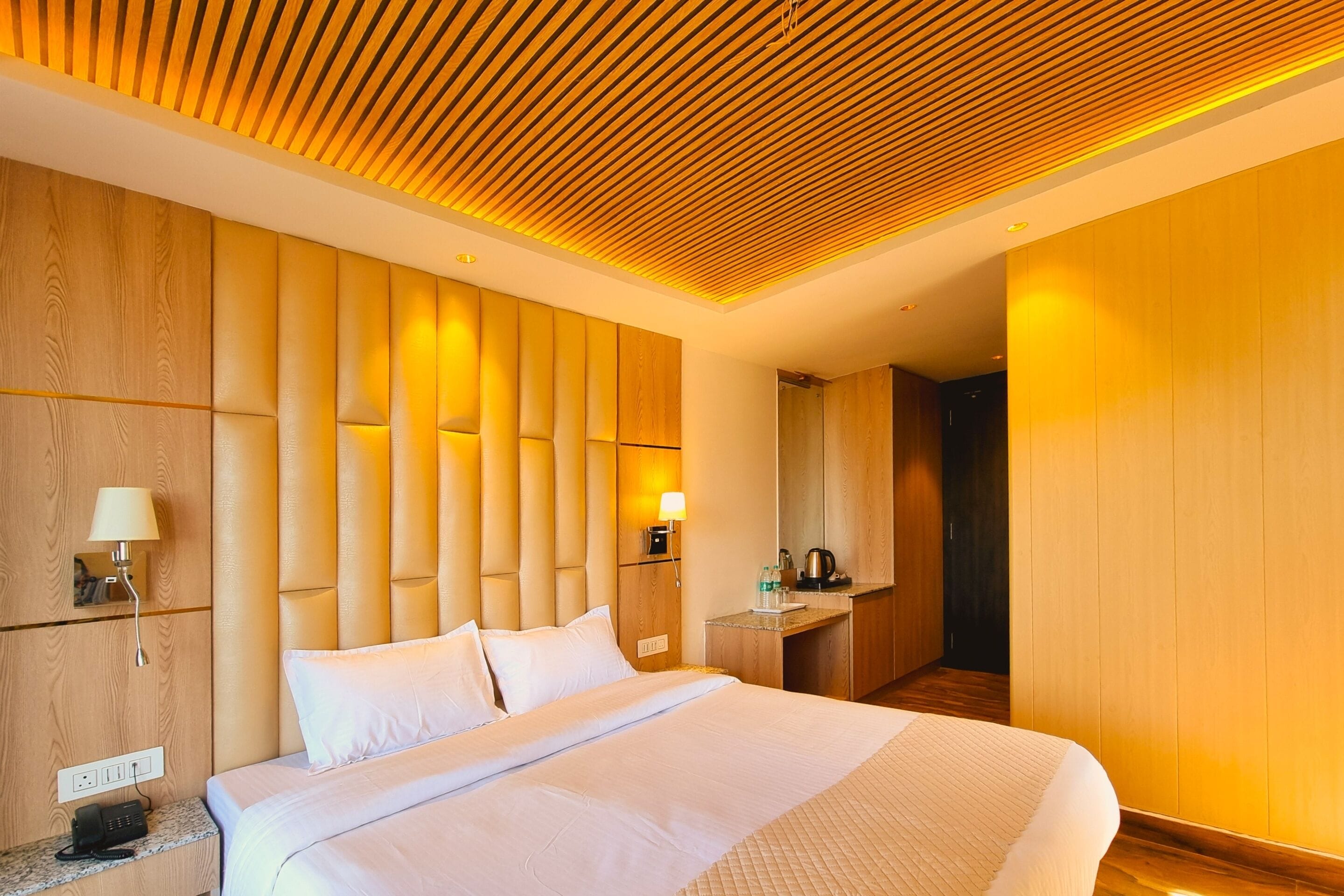 Una habitación de hotel moderna con una cama grande, cabecera de madera y una pequeña zona de escritorio con lámpara y teléfono.