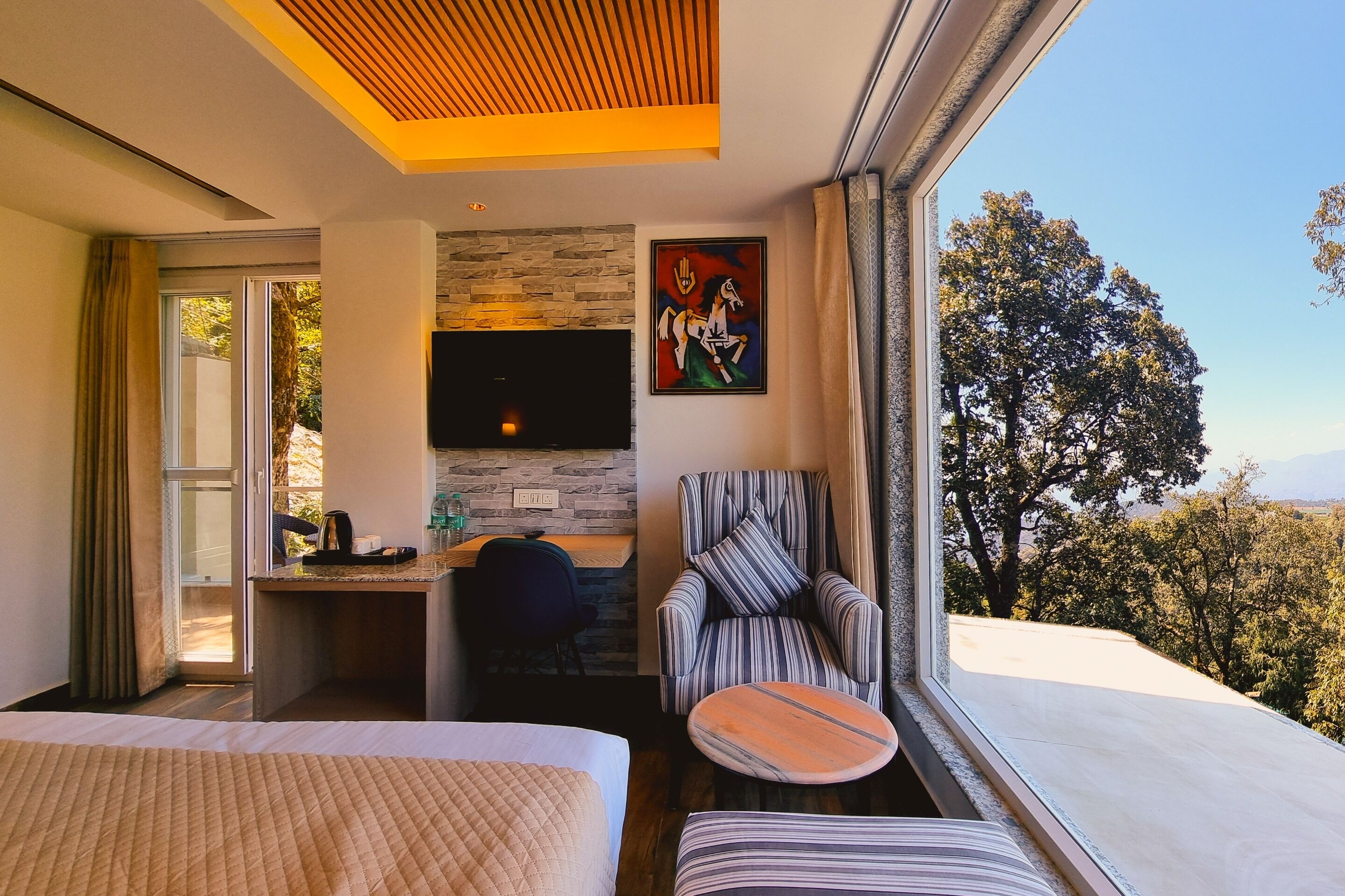 A hotel room with a bed, a desk with a chair, a television, and a view of trees.